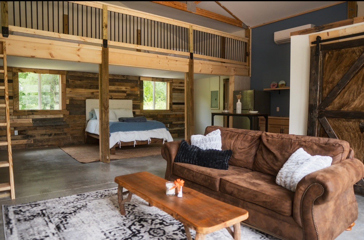 An inviting living area is highlighted by a plush brown sofa and a wooden coffee table. Natural light enters through large windows, illuminating the space. Above, a sleeping area is visible with a spacious bed, surrounded by rustic wooden accents.