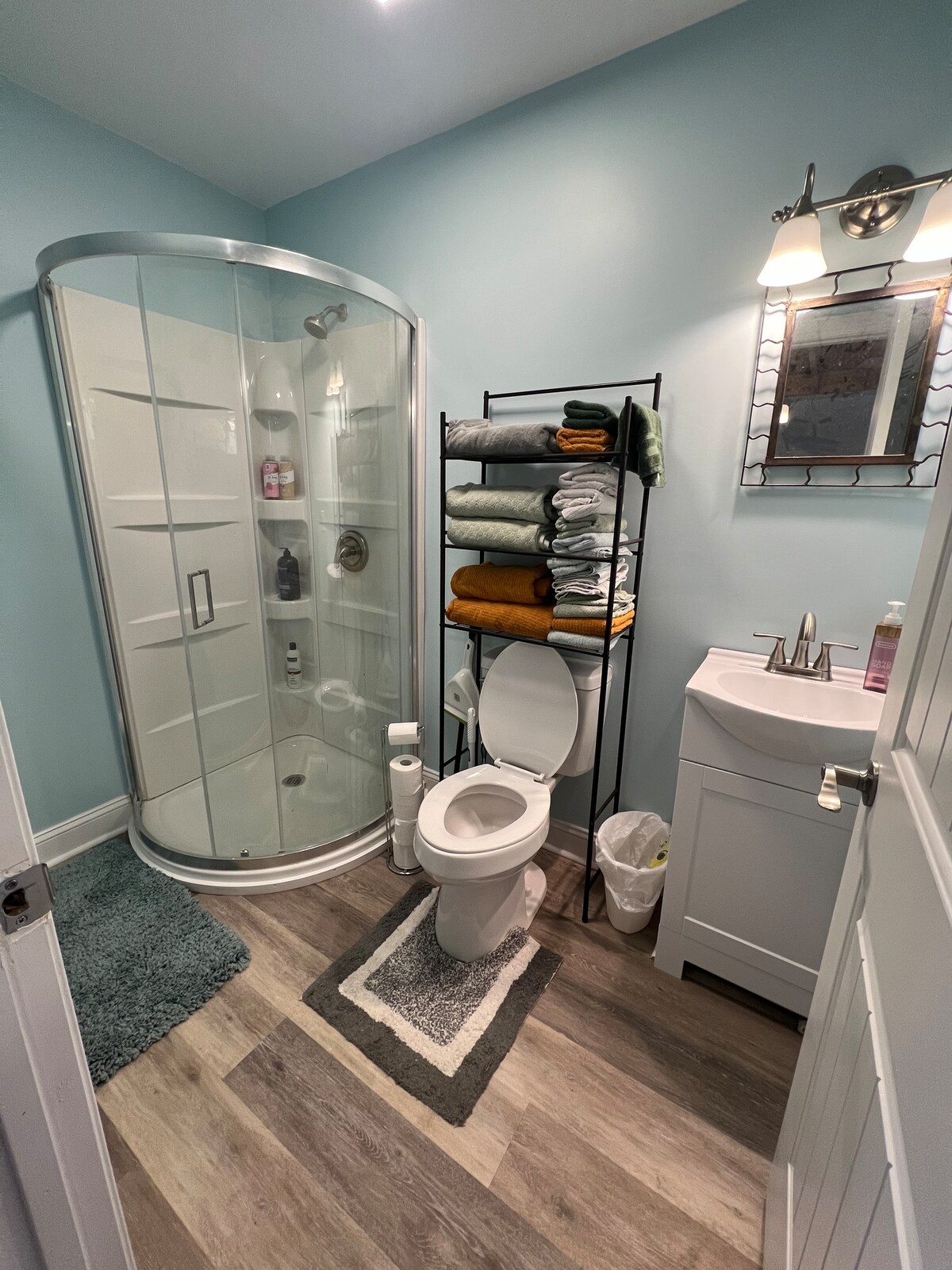A compact bathroom features a glass-enclosed shower with modern fixtures. Soft towels are neatly stacked on a metal shelf beside the toilet. A small sink with a mirror and a wastebasket are included, with light blue walls enhancing the clean appearance.