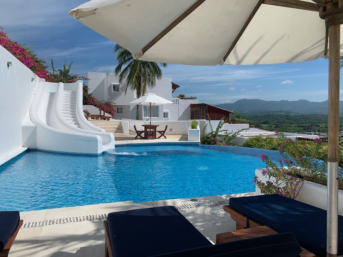 A spacious pool with smooth, flowing slides is surrounded by lush tropical greenery. Sun loungers are positioned nearby under a large umbrella, inviting relaxation. The two-story villa can be seen in the background, complementing the scenic mountain view under a clear blue sky.