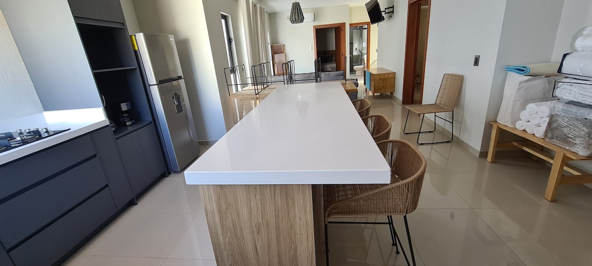 A modern kitchen space features a large white island countertop surrounded by woven bar chairs. Stainless steel appliances, including a fridge, are present, and a flat-screen TV is mounted on the wall. Ample natural light filters through large windows.