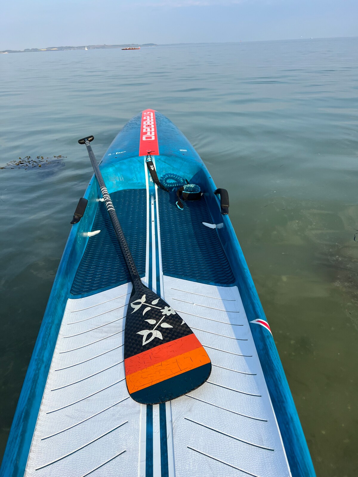 A paddle board is displayed on calm, clear water, showcasing its striking blue surface highlighted by a textured design. A paddle rests across the board, its vibrant colors reflecting a sense of adventure. Nearby, subtle ripples suggest a peaceful atmosphere on the water.