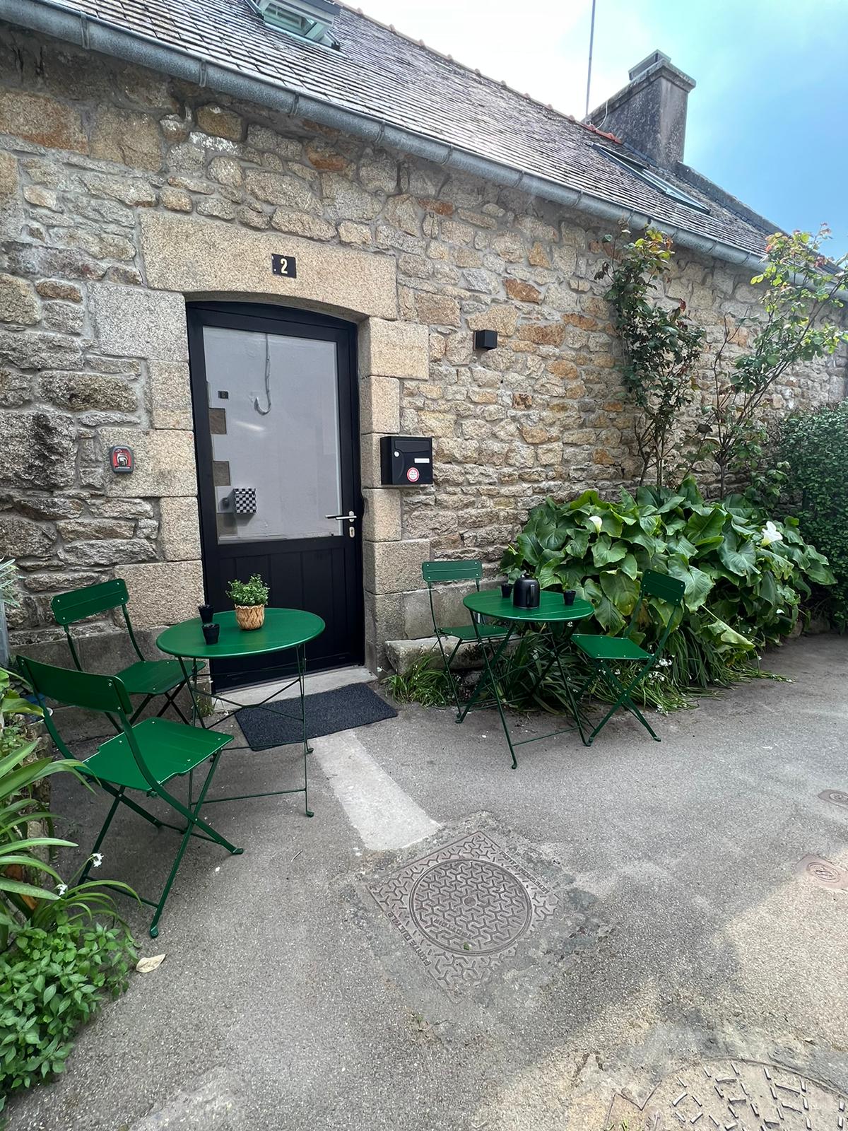 A charming outdoor area is presented, featuring two round green bistro tables with matching chairs and a small potted plant. The stone facade of the building adds rustic character, while lush greenery frames the entrance, creating a serene and inviting ambiance.