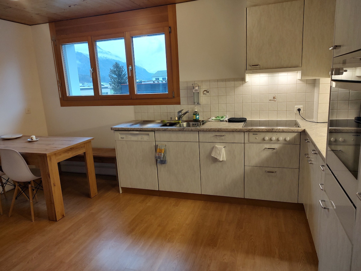 A functional kitchen area features light wood cabinetry and a spacious countertop. A dining table made of natural wood is present, accompanied by modern white chairs. Large windows provide mountain views and natural light, enhancing the overall brightness of the space.