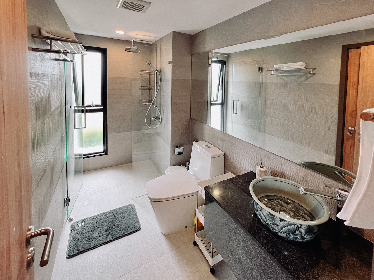 A modern bathroom is featured, showcasing a sleek design with a glass walk-in shower and a contemporary toilet. A stylish vanity with a unique basin is accompanied by dark granite countertops, and natural light filters in through an adjacent window, enhancing the space's openness.