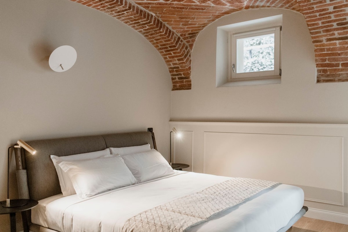 A spacious bedroom features a comfortable bed with a neutral-colored bedspread. Soft lighting is provided by bedside lamps, and a small window allows natural light to illuminate the room. The arched brick ceiling adds architectural interest to the welcoming environment.