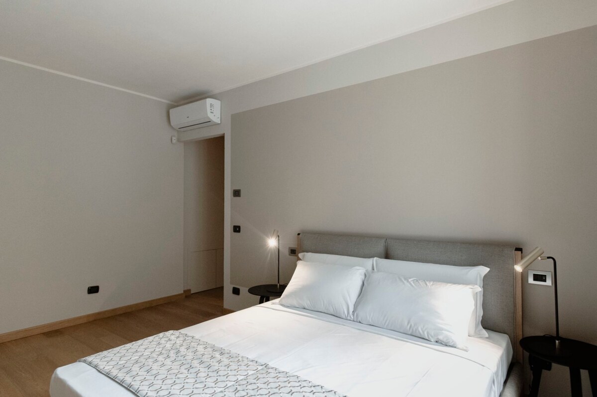 The airy bedroom features a spacious bed dressed in crisp white linens, flanked by modern nightstands with reading lights. A soft neutral color palette creates a calming ambiance. A wall-mounted air conditioning unit is positioned for comfort, providing a functional yet stylish touch to the space.