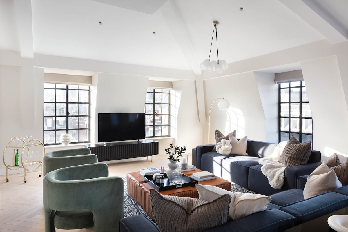 A spacious living area showcases large crittal windows allowing natural light to fill the room. Comfortable seating is arranged around a central coffee table, accentuated by soft pillows and throws. A modern television is mounted on the wall, contributing to a cohesive, inviting atmosphere.