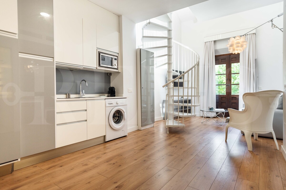 The open-plan lower level features a fully equipped kitchen with modern appliances, including a washing machine. A spiral staircase leads to the upper floor. Natural light floods the space from a large window, which frames views of greenery outside. A comfortable chair is positioned near the entrance.