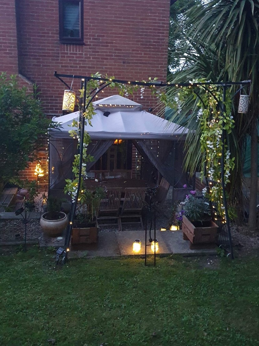 A cozy outdoor seating area is enclosed by soft fabric with twinkling lights. The space features wooden furniture surrounded by potted plants, creating a serene retreat. Lanterns add gentle illumination to the pathway leading to the seating.