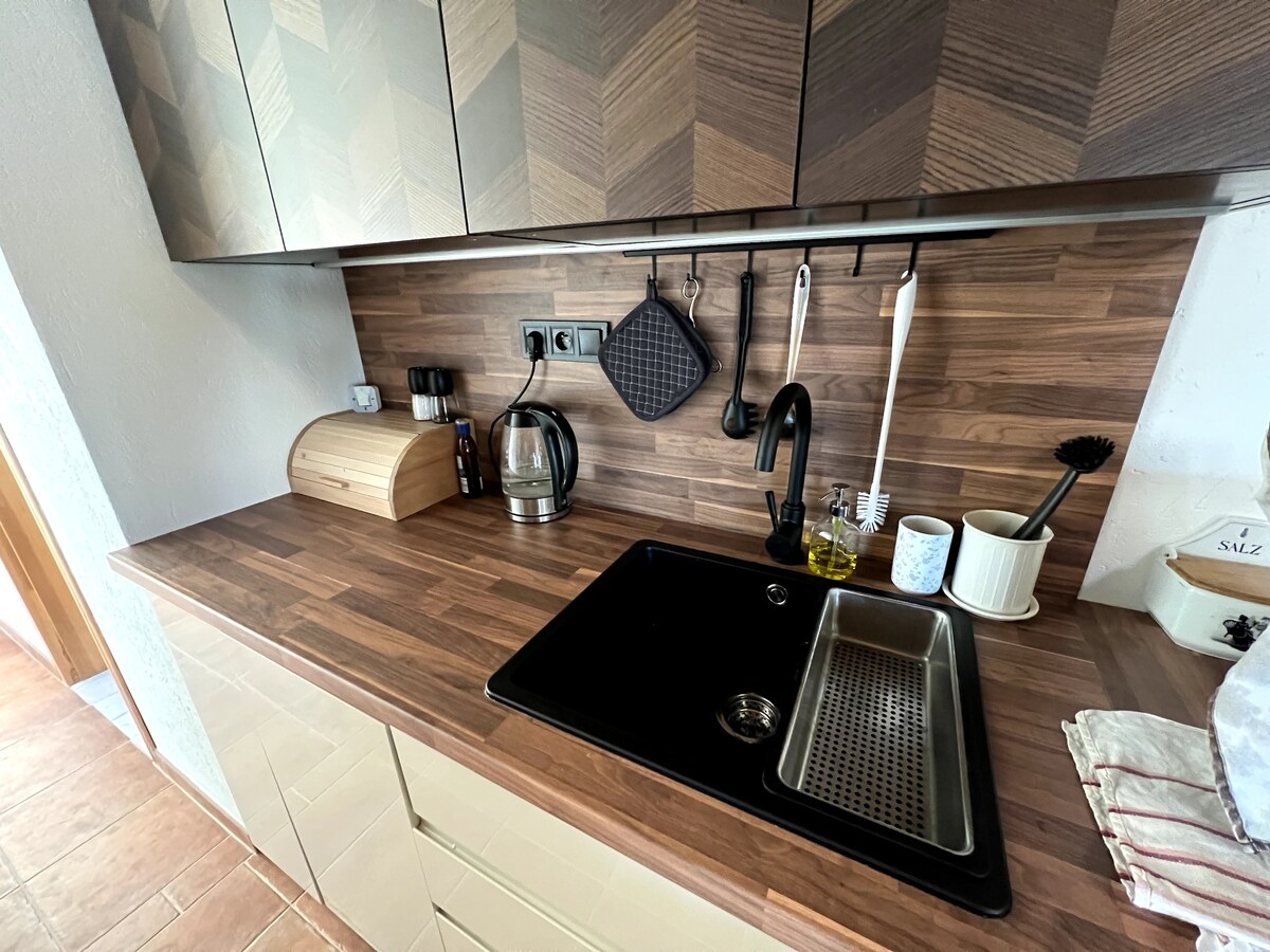 A well-equipped kitchen features a wooden countertop with a sink and a kettle. Kitchen utensils are organized on a hanging rack, complemented by a wooden breadbox. A modern black faucet stands above the sink, emphasizing functionality and convenience.