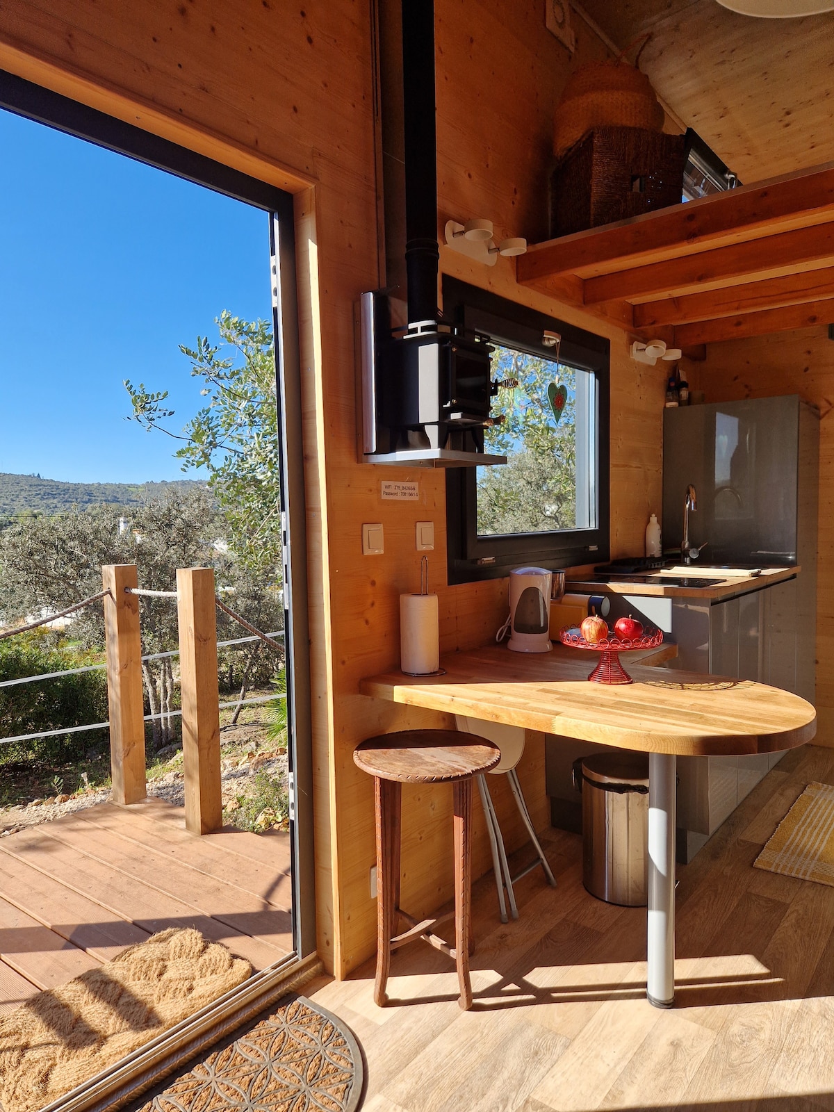 A wooden interior features a small dining table set with a fruit bowl. A modern kitchenette is equipped with essential appliances. Large glass doors open to a scenic view, allowing natural light to brighten the space. The warm wood finishes create a cozy atmosphere.