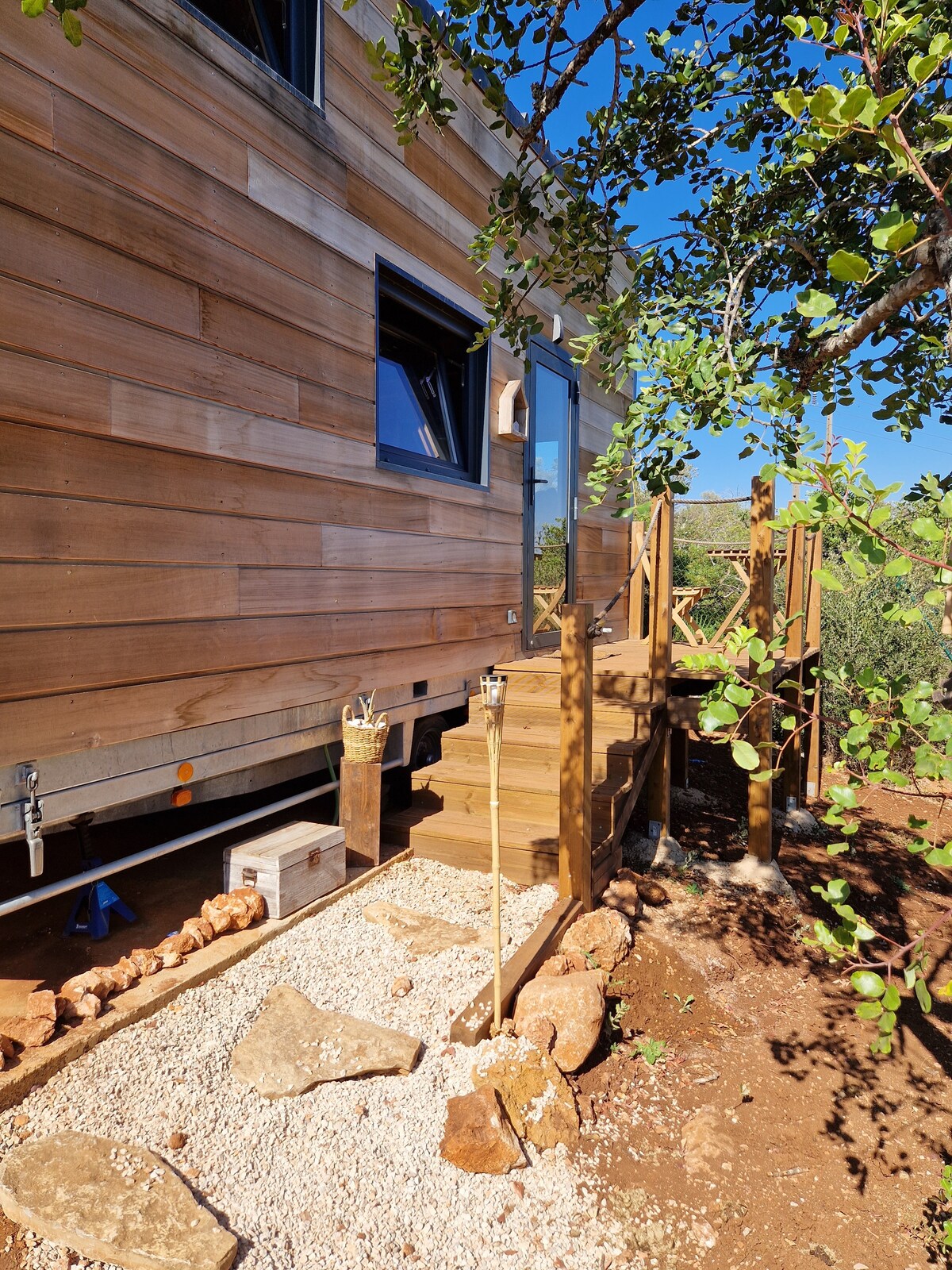 A wooden exterior of a tiny house is presented, featuring a series of steps leading to the entrance. Decorative stones and a gravel pathway appear alongside lush greenery, enhancing the natural setting. A clear sky is visible above, contributing to a serene outdoor environment.