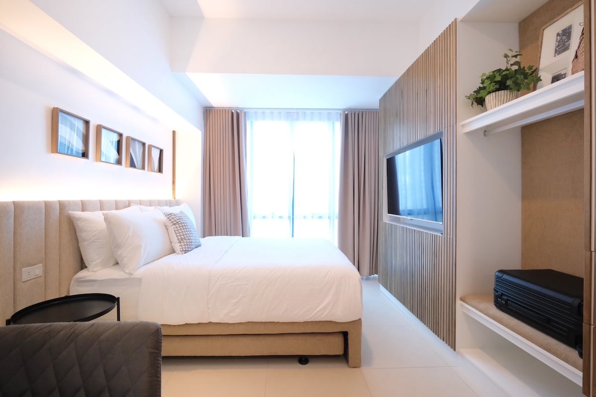 A well-lit studio space features a queen-sized bed with a cushioned headboard and crisp white linens. A modern flat-screen TV is mounted on the wooden accent wall, while large windows allow natural light to brighten the room. A small luggage rack is positioned by the shelves.
