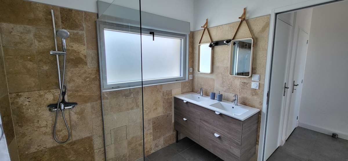 A modern bathroom is presented, featuring a large walk-in shower and dual sinks positioned on a wooden vanity. Neutral stone walls complement the space, while a mirror and minimalist decor enhance the overall design. Natural light filters in through a frosted window.