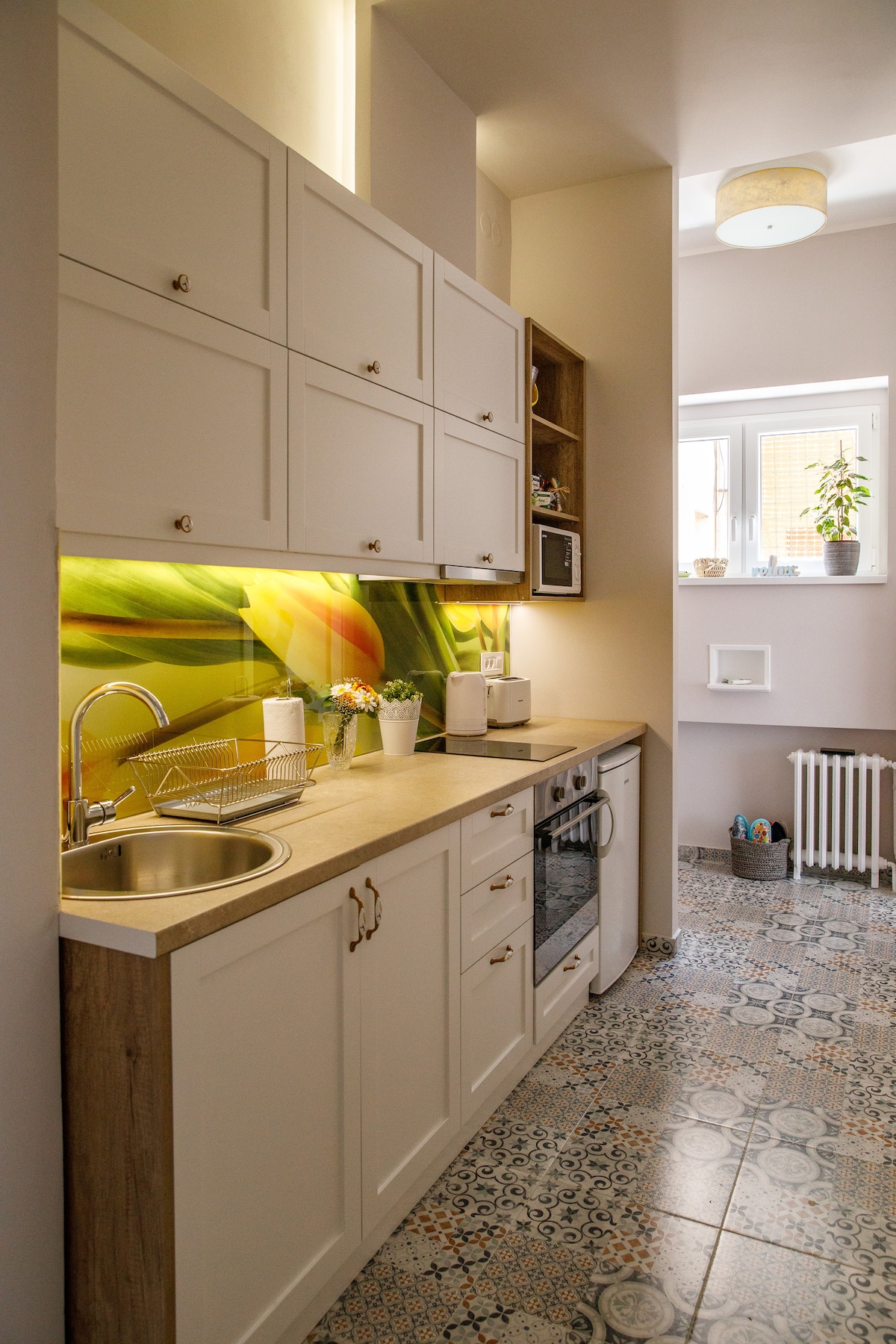 A compact kitchen space is equipped with modern appliances, including an oven and a sink. Light-colored cabinetry is paired with a decorative backsplash featuring floral imagery. Natural light enters through a small window, illuminating the patterned floor tiles.