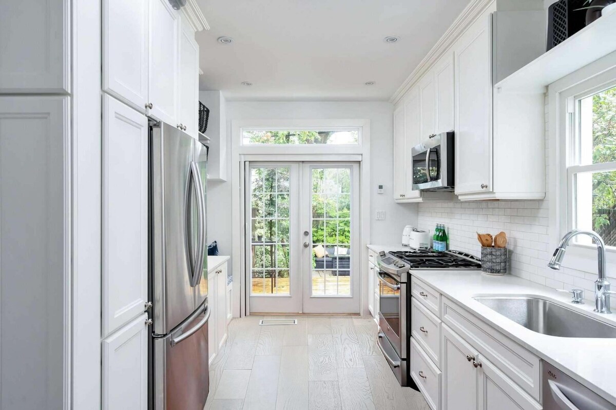 The kitchen features white cabinetry and stainless steel appliances, including a refrigerator and microwave. A double door leads to an outdoor area, while a spacious sink is situated under a window, allowing natural light to illuminate the space.