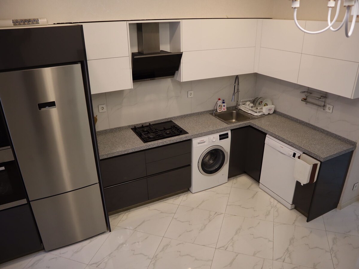 The kitchen features modern cabinetry in a sleek black and white design. A stainless steel refrigerator, washing machine, and dishwasher are included. A gas stove and sink are positioned against a spacious countertop, with marble flooring adding a touch of elegance to the space.