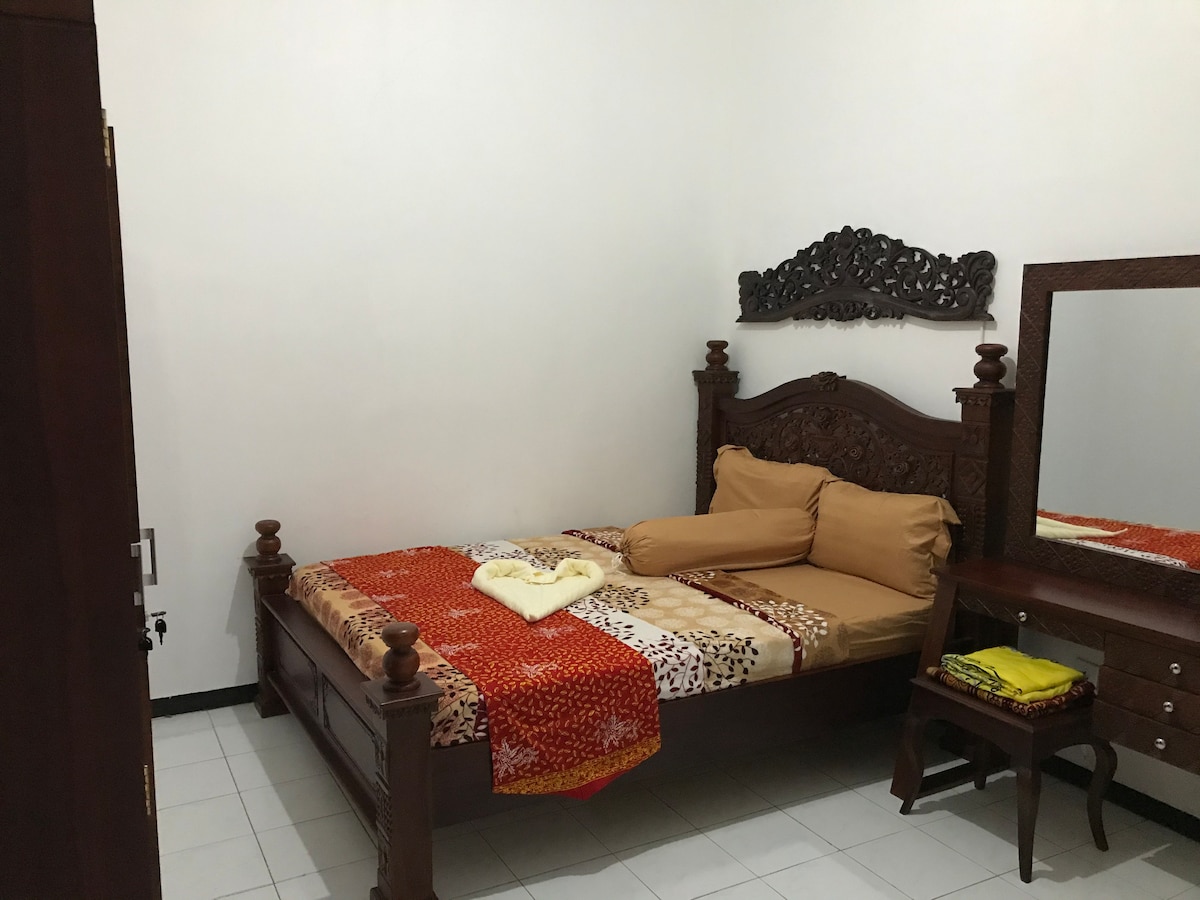 A traditional-style bedroom features a wooden bed with ornate detailing and matching bedside furniture. Soft bedding is layered in warm tones, complemented by a decorative mirror on the wall. The room is illuminated by natural light filtering through the space.