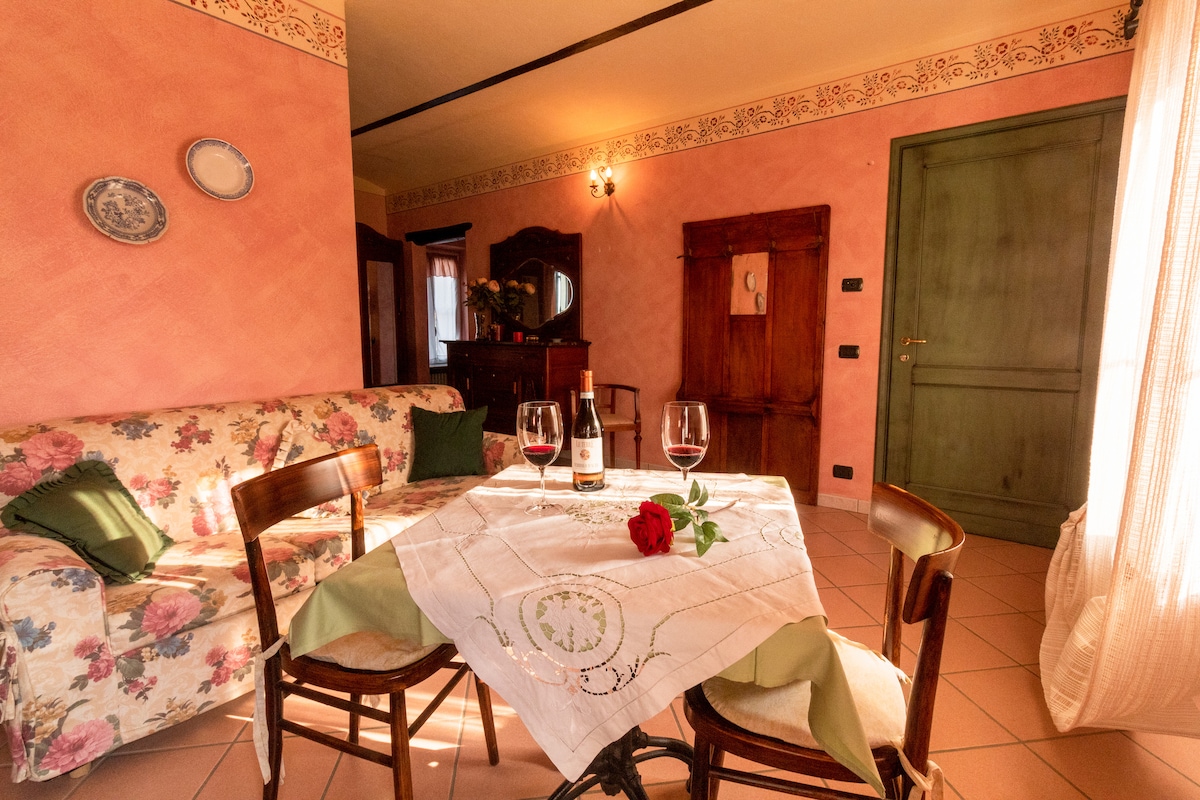 A dining area is presented with a small table adorned with a white tablecloth and a single red rose. Surrounding chairs complement a floral-patterned sofa. The soft pink walls create a warm ambiance, while natural light filters through a nearby window, highlighting the space.