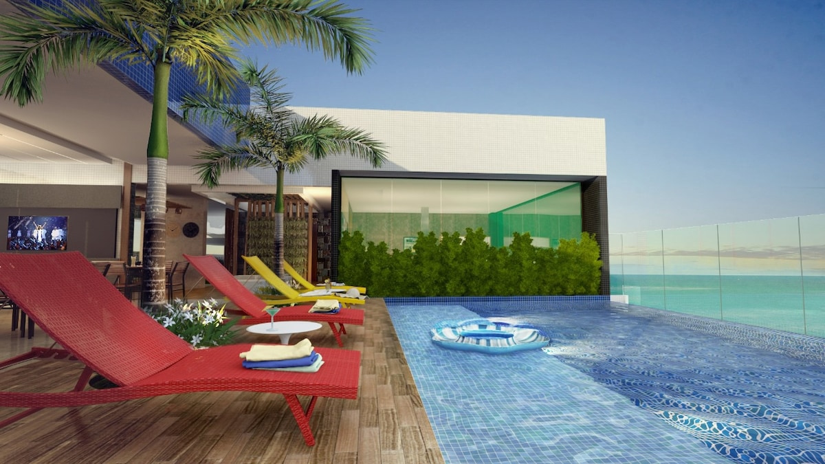 An outdoor pool area features a panoramic view of the ocean. Colorful loungers in red and yellow are accompanied by small side tables. Lush greenery provides a natural backdrop against the modern architecture of the building.