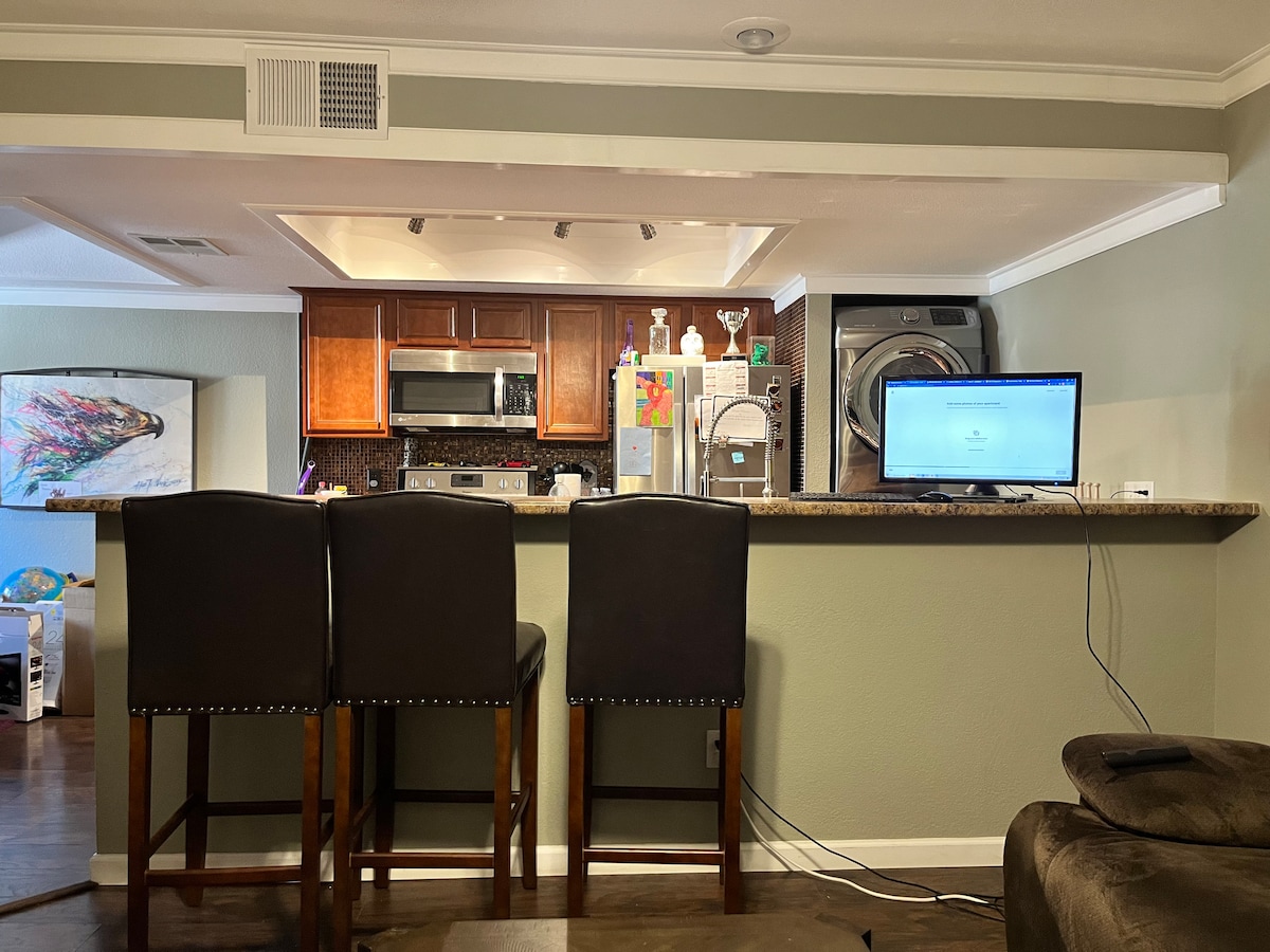 A modern kitchen is visible, featuring dark wood cabinetry and stainless steel appliances. Three high stools are positioned at a granite countertop. A computer screen is placed on the counter, with a washing machine and dryer situated in the background.