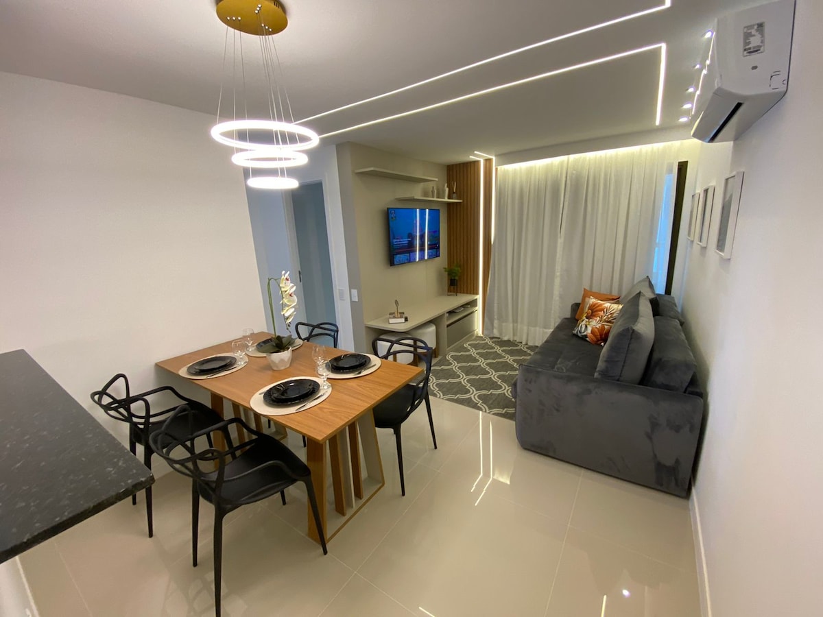 An open living space features a dining table with four black chairs, complementing a comfortable gray sofa. A smart TV is mounted on the wall, and soft lighting enhances the modern aesthetic. Light-colored curtains partially obscure the windows, allowing natural light to fill the area.