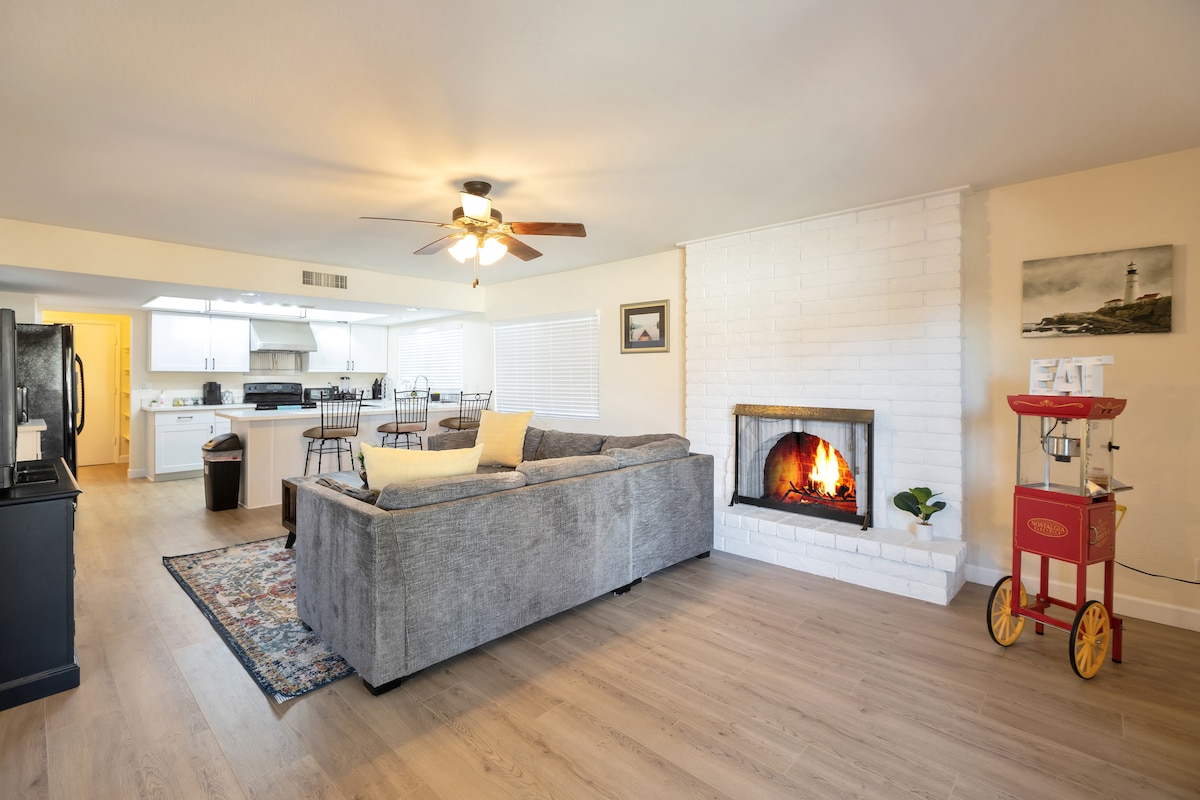 A spacious living area features a comfortable gray sectional sofa, complemented by a fireplace with a crackling fire. In the background, the open kitchen is visible, showcasing modern appliances and a dining space with black chairs and a table. Natural light enters from the window.