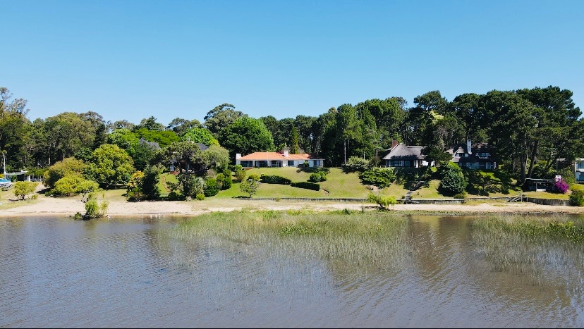 A serene landscape features a spacious house set on a lush, green lawn by the water's edge. Trees and colorful shrubs surround the property, while a calm lagoon reflects clear blue skies. The sandy beach area offers a natural transition to the water.