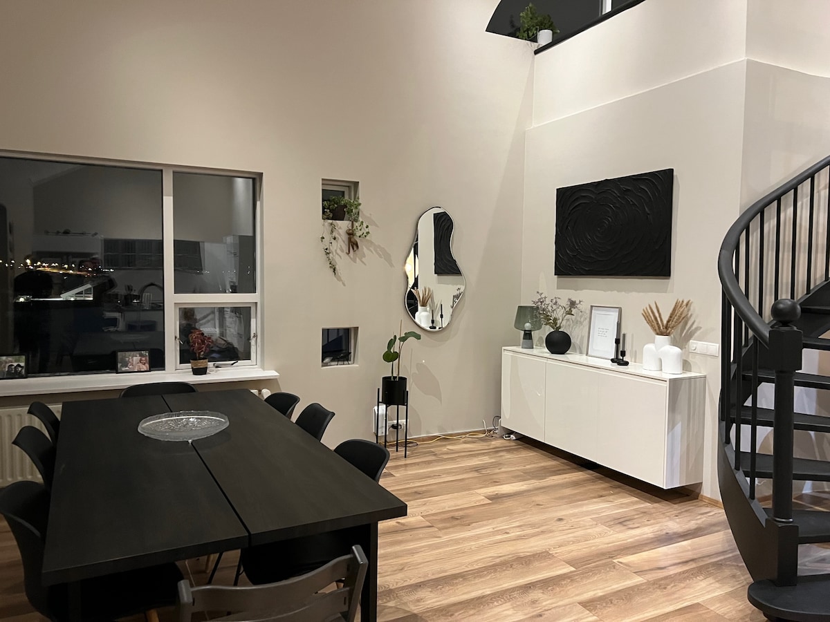A modern dining area features a large black table surrounded by dark chairs. Natural wood flooring complements the neutral walls, while a large mirror reflects light and space. A decorative wall piece adds texture, and potted plants are placed strategically throughout the room.