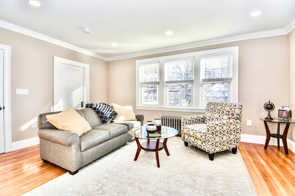 A comfortable living area is illuminated by natural light from the windows. A gray sofa is accented with bright throw pillows, while a round glass coffee table is placed in front. A patterned armchair complements the space, adding a touch of style.