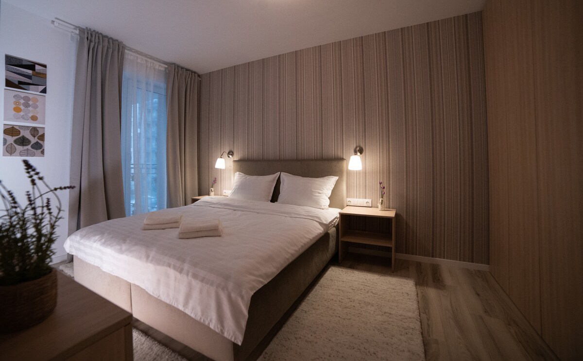 The bedroom features a spacious bed with crisp white linens, flanked by two bedside lamps. A subtle striped wall adds warmth, and natural light filters through sheer curtains. A small plant is placed on the nightstand, contributing to the calming atmosphere.