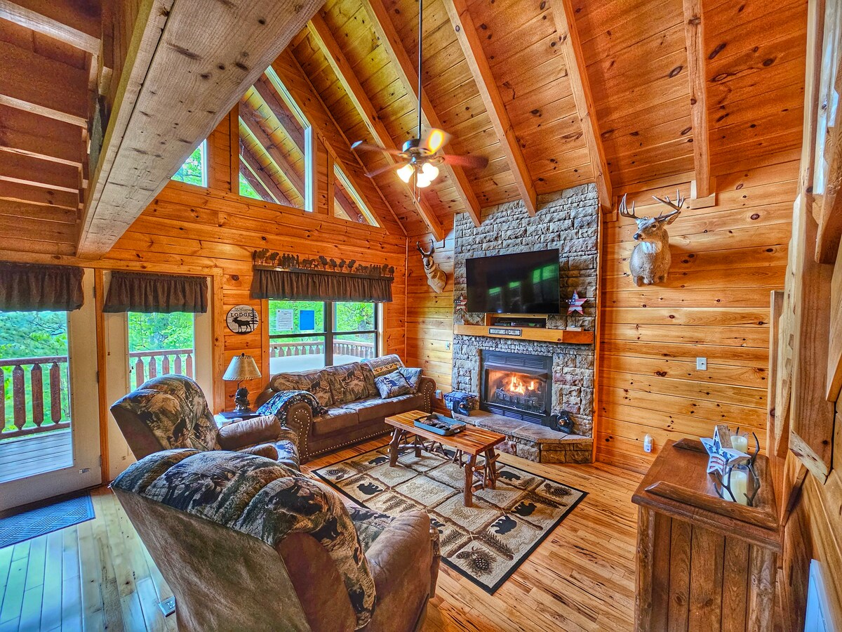 The living room features a high, wooden beam ceiling with large windows allowing natural light. A stone fireplace with a gas log fire is centered against a rustic wall, complemented by comfortable seating. Decor includes a patterned area rug and wall-mounted decor, creating a cozy environment.