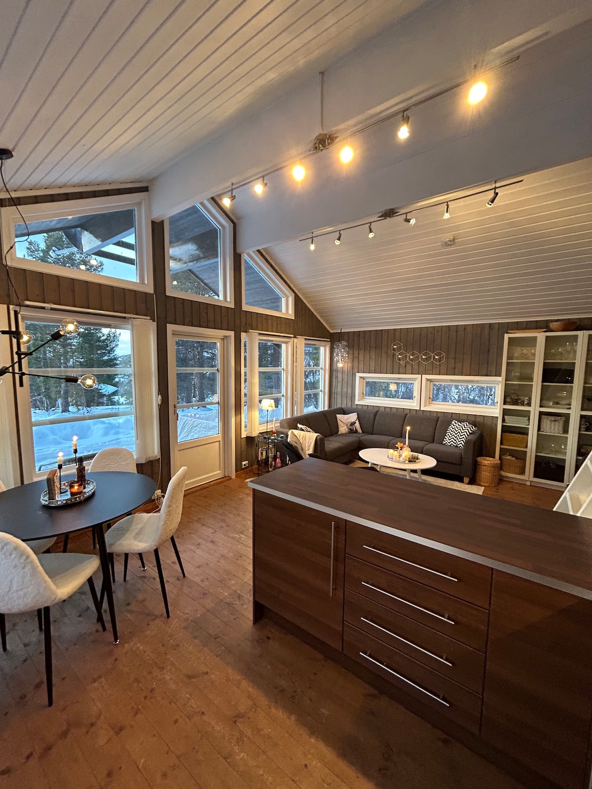 An open-plan living space is highlighted by large windows that allow natural light to fill the area. A modern sofa is positioned beside a dining table. The kitchen island features sleek cabinetry. Light fixtures add warmth to the wood-paneled walls.