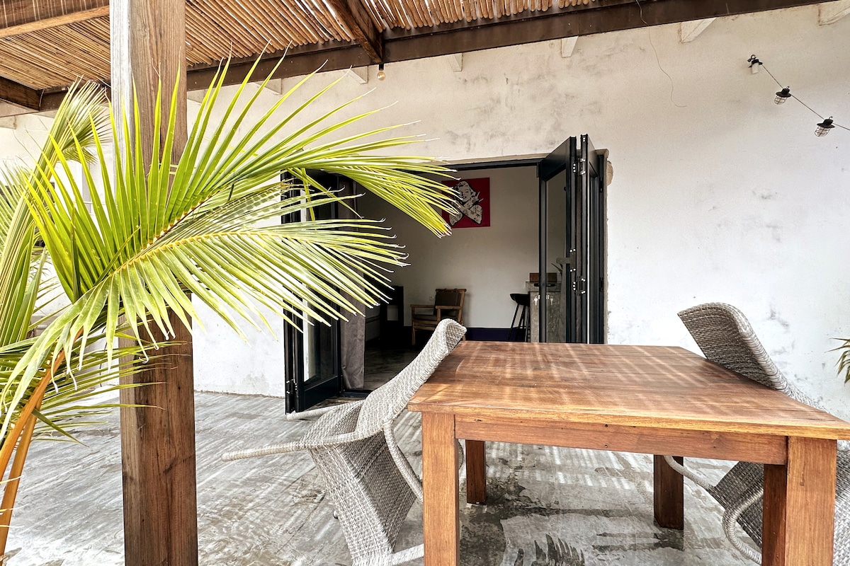 An inviting outdoor space features a wooden table surrounded by two woven chairs. A palm frond adds a touch of greenery while large sliding doors open into the interior, allowing natural light to fill the area.