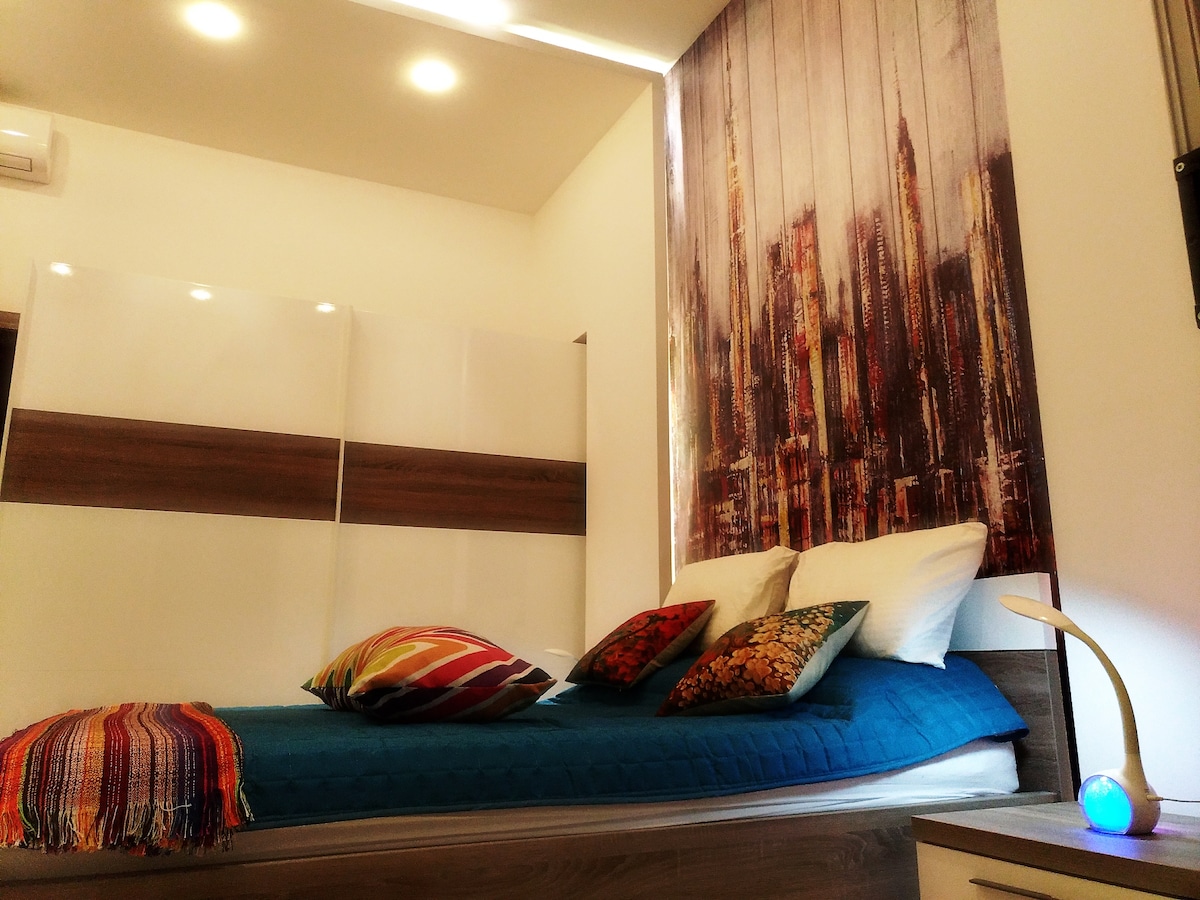 A bright sleeping area features a spacious bed adorned with colorful pillows. A vibrant throw blanket accents the bed, while a modern light fixture provides illumination. A textured wall design depicting an urban skyline enhances the contemporary decor.