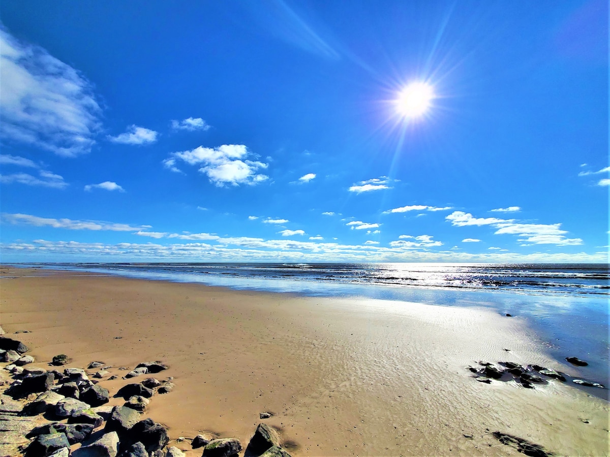 A wide expanse of beach stretches towards the horizon under a clear blue sky adorned with a bright sun. Gentle waves reflect shimmering sunlight, while smooth rocks line the edge of the sandy shore.