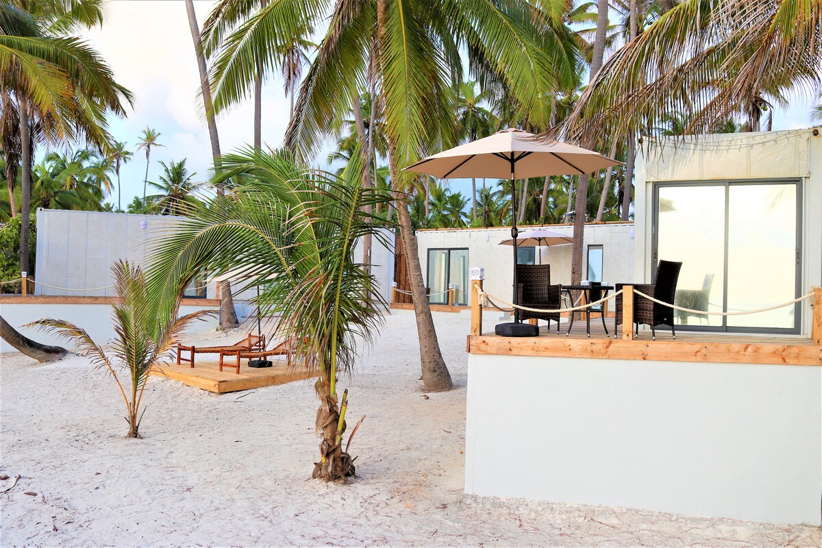 A beach-front deck features comfortable wicker chairs and a table under a sun umbrella, surrounded by soft white sand and palm trees. In the background, mini villas are visible, providing a serene coastal environment.