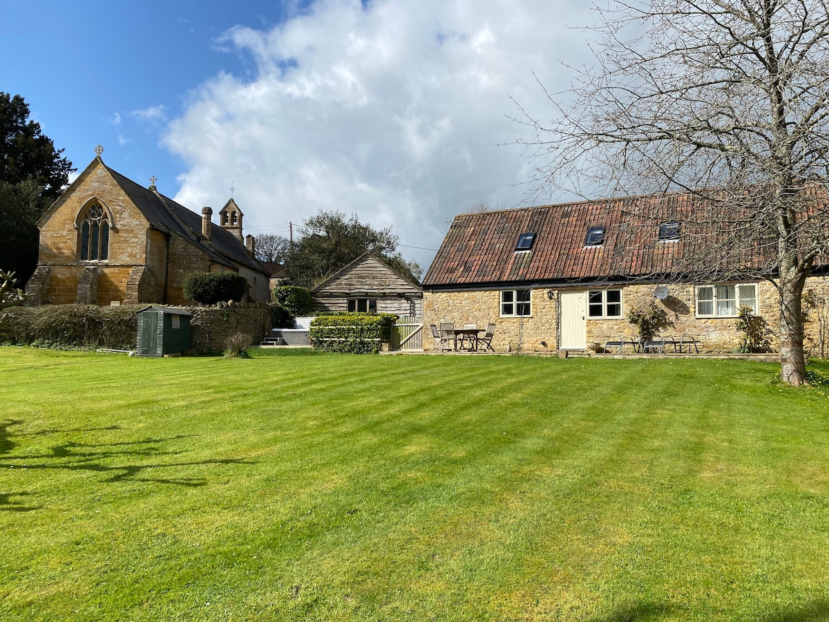 A spacious green lawn surrounds the cottage, showcasing a well-maintained landscape. The building features a rustic charm, with a red-tiled roof and multiple windows. Nearby, an architectural structure with pointed arches adds character to the setting, creating a peaceful rural atmosphere.