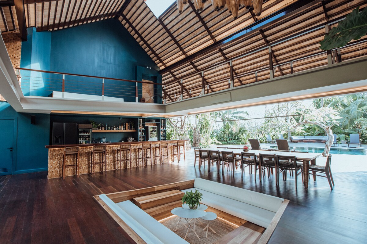 An open-concept living space features high ceilings with wooden beams, showcasing a modern bar area with seating. Natural light floods the room, highlighting a sunken lounge area surrounded by a wooden platform. Outdoor views of lush greenery and a pool create a serene environment.
