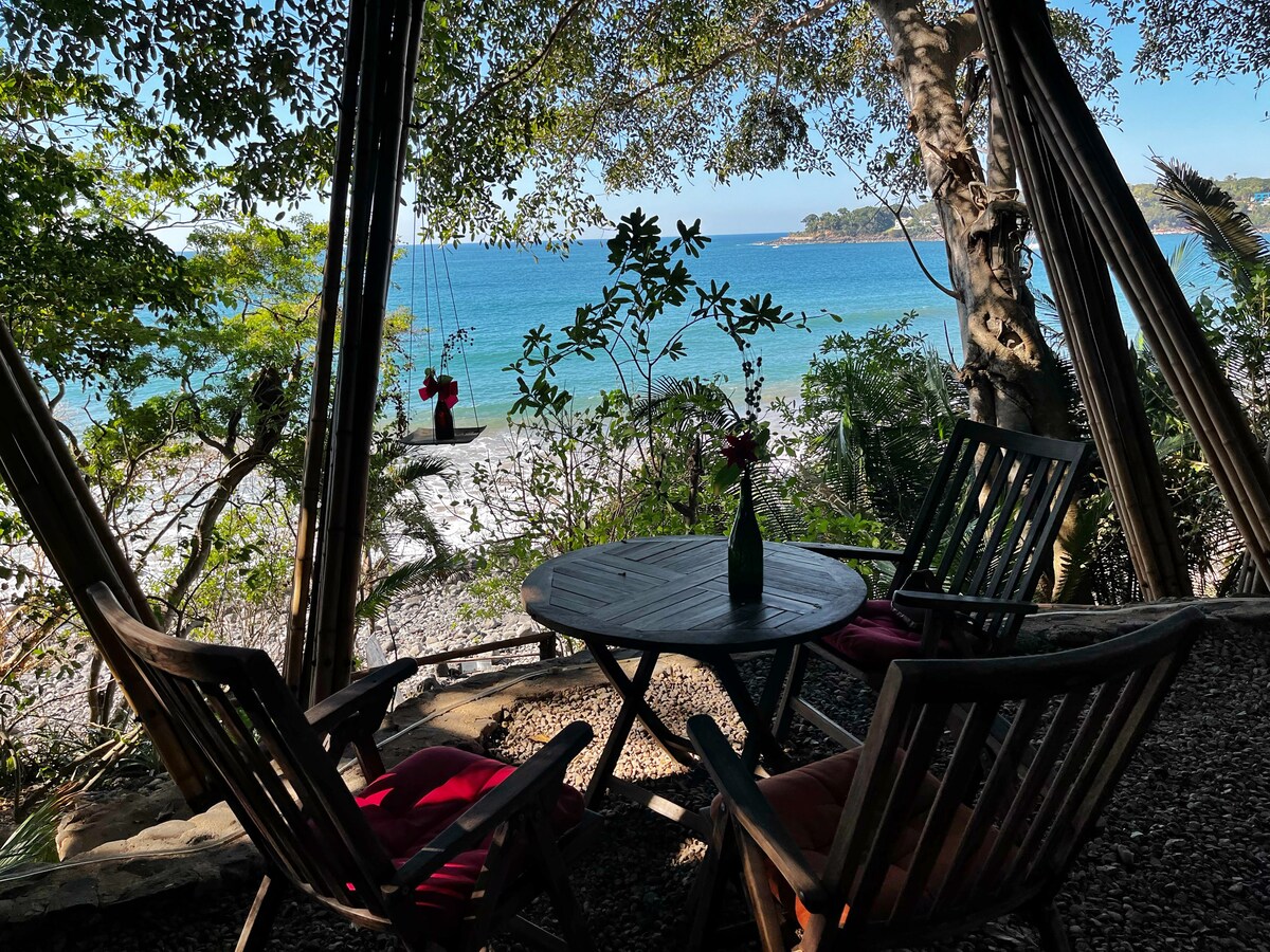 A tranquil outdoor seating area is surrounded by lush vegetation. Two wooden chairs with cushions and a small round table are visible. In the background, the blue water of the sea can be seen through the trees, with a swing hanging nearby.