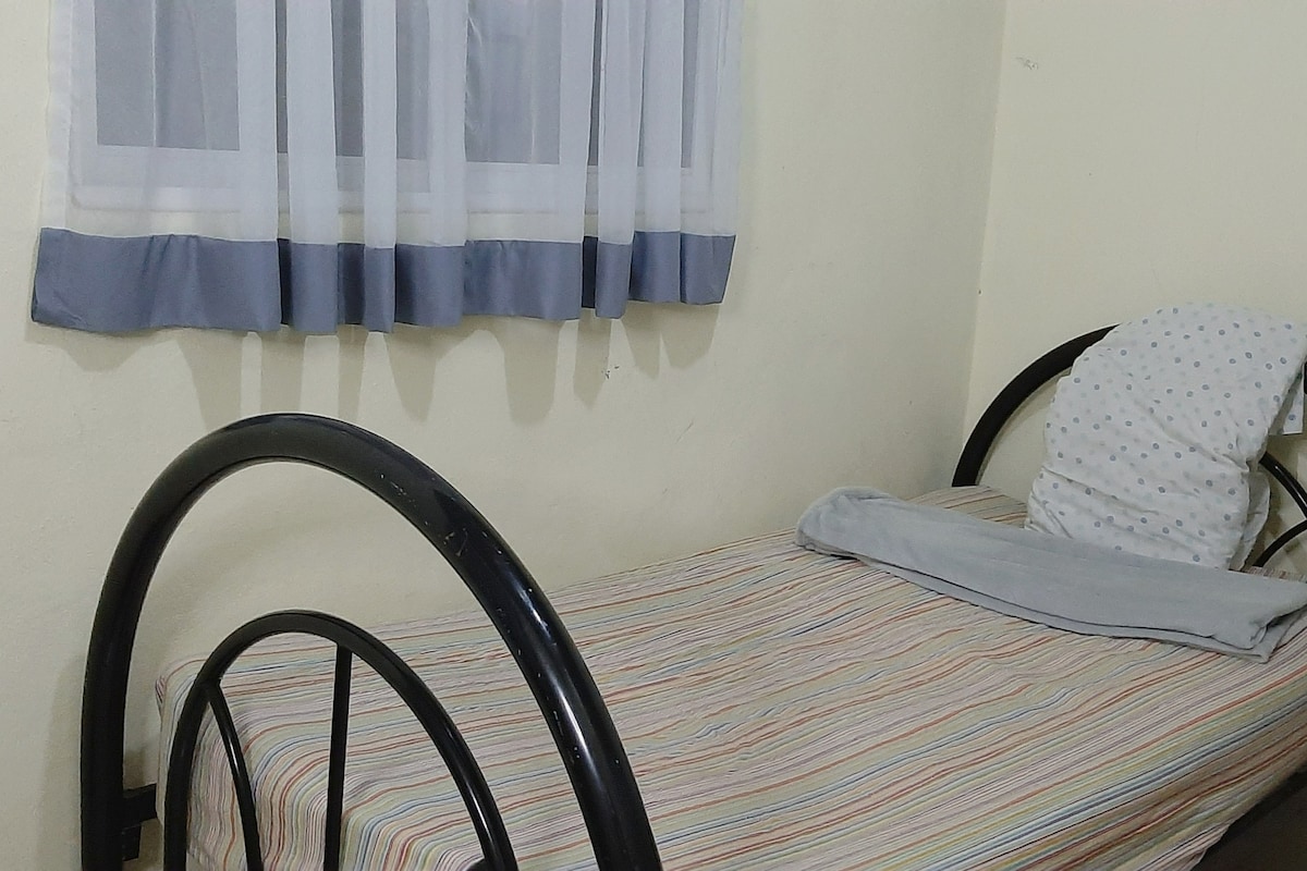 A single bed features a striped bedspread and a light grey blanket. A neatly arranged pillow is placed against the wall’s corner. Sheer white curtains with grey trim frame a window, allowing natural light to brighten the cozy room.