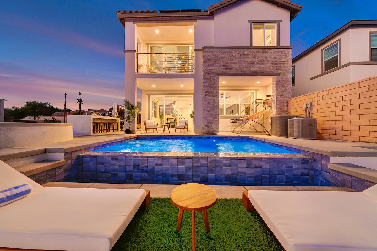 An inviting outdoor area features a shimmering blue pool surrounded by well-maintained grass. Two loungers provide comfortable seating, while a small wooden table is situated nearby. The back of the house showcases large windows and a balcony, merging indoor activities with the serene outdoor environment.
