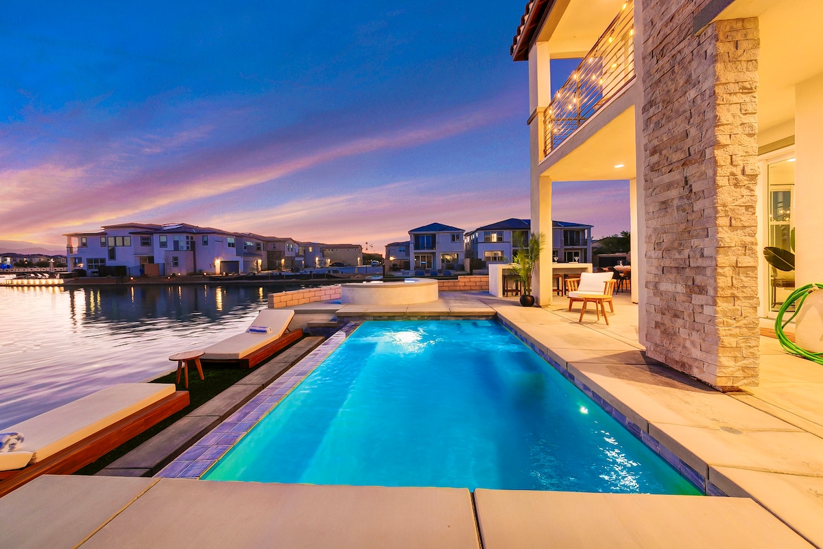 A serene outdoor space is presented, featuring a sparkling pool alongside a tranquil lake. Comfortable lounge chairs are positioned nearby, with warm sunset hues reflecting off the water's surface. The backdrop showcases interconnected homes along the water's edge, enhancing the peaceful atmosphere.