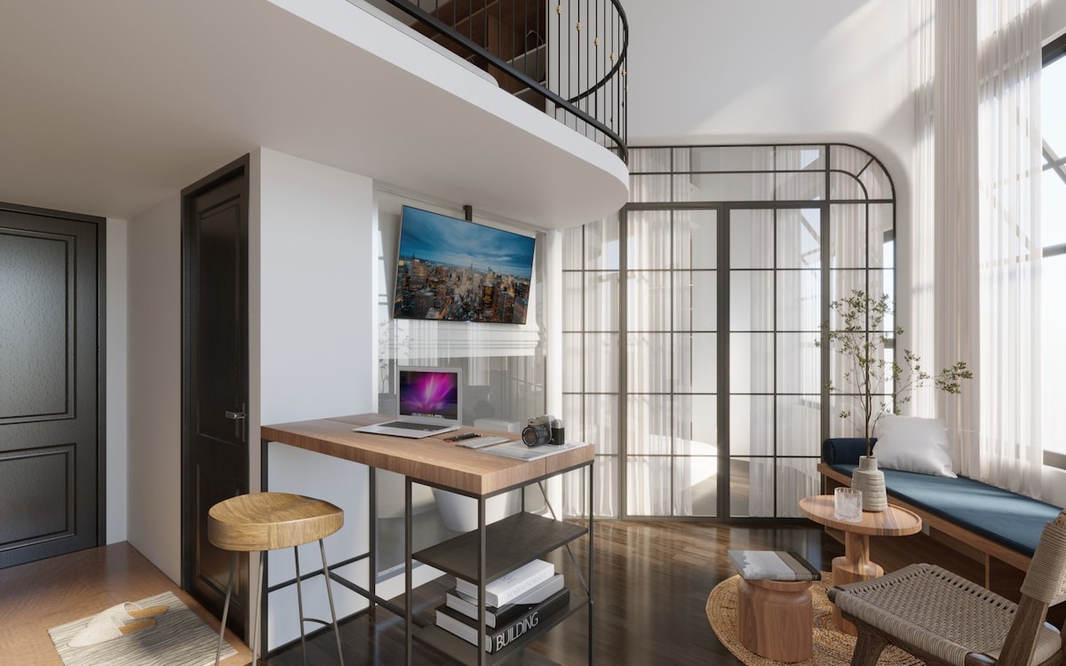 A modern living area showcases a stylish table supported by metal legs, accompanied by two high stools. A large screen and a laptop are arranged on the table, while natural light flows through large windows. A comfortable seating area features a sofa and a small round table.