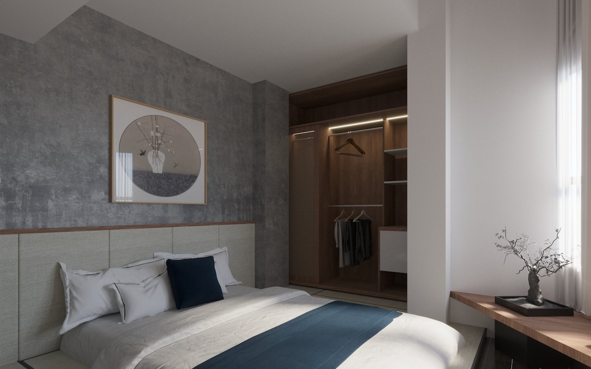 A serene bedroom features a spacious bed draped in soft linens, with a decorative blue throw positioned at the foot. A wooden wardrobe stands in the corner, illuminated from within, and a minimalist nightstand holds a simple plant arrangement.