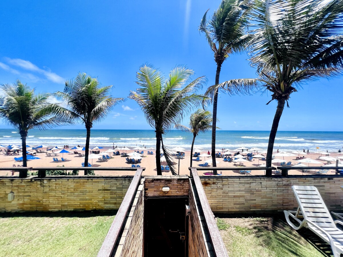 A scenic view is presented from a patio, featuring palm trees in the foreground and a sandy beach with umbrellas in the distance. The ocean stretches out under a clear blue sky, inviting relaxation and enjoyment of the coastal atmosphere.