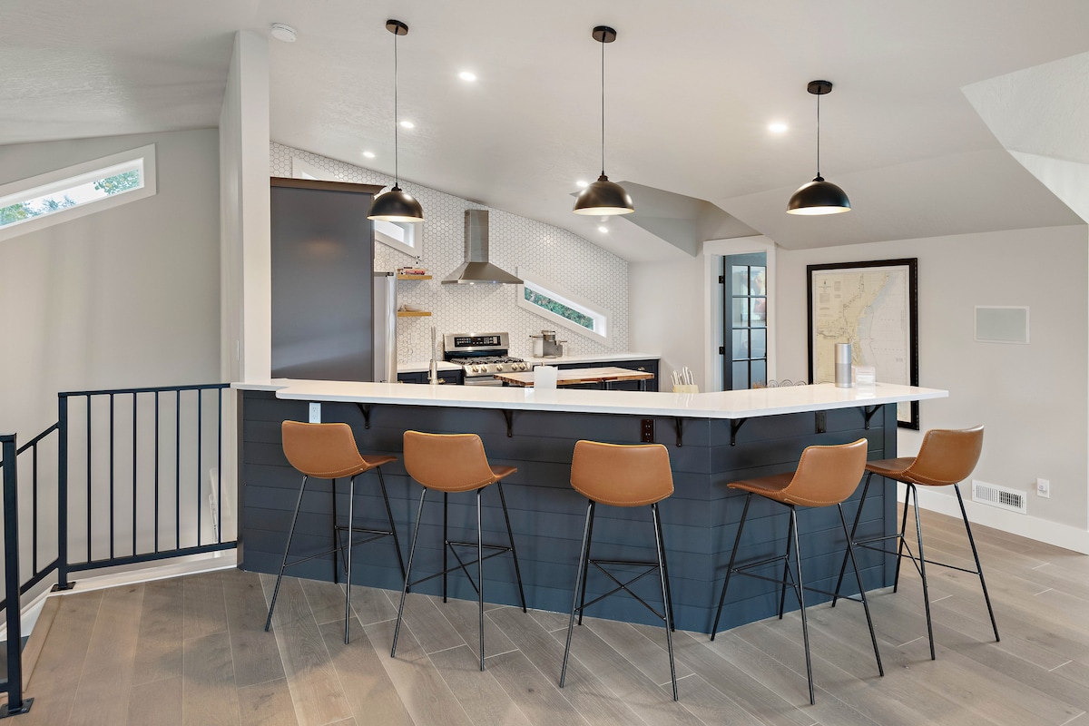 A modern kitchen island features seating for four with tan bar stools. Pendant lighting hangs above a white countertop, while stainless steel appliances and geometric backsplash tiles provide a clean and stylish cooking space. Large windows allow natural light to fill the area.