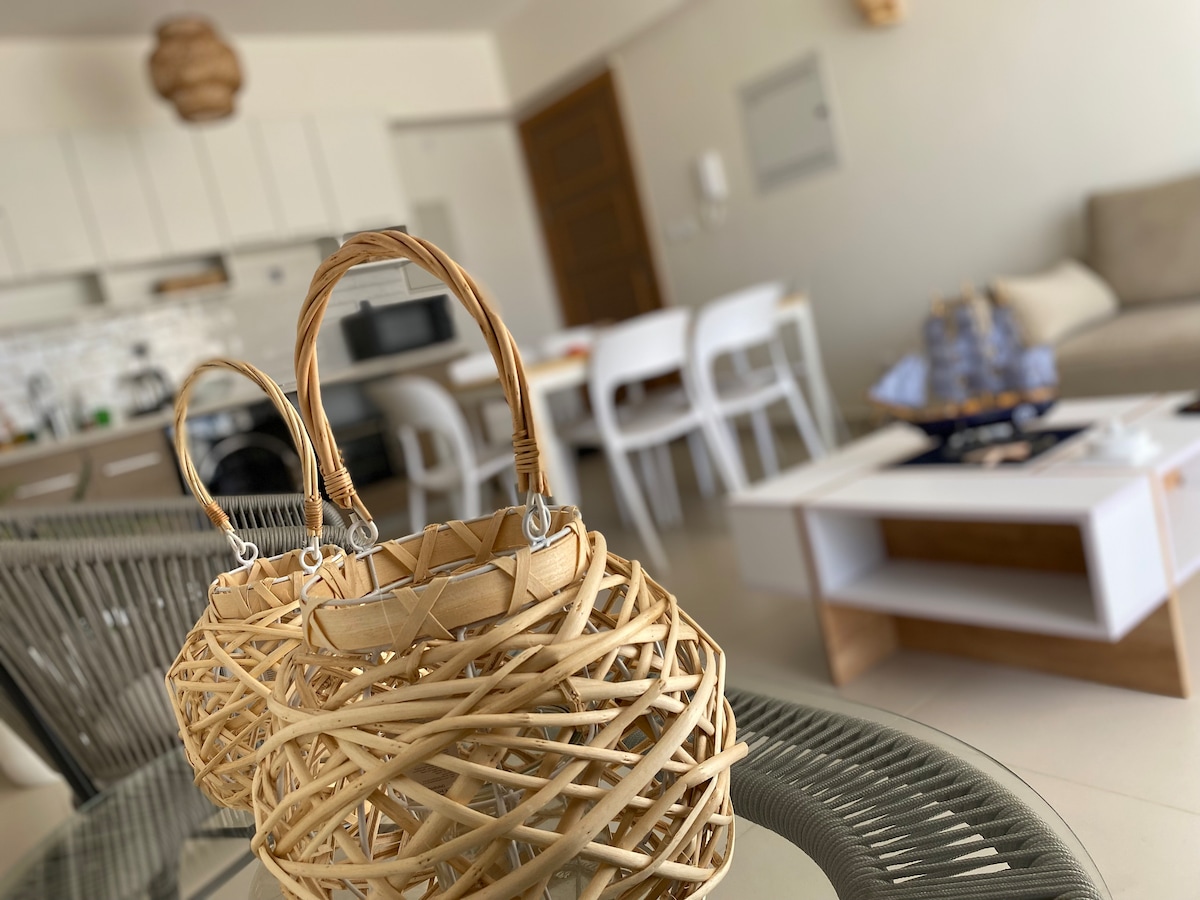 Woven baskets are positioned in the foreground, showcasing intricate detailing. In the background, a modern kitchen is visible, equipped with sleek appliances and a light-colored dining set. A comfortable seating area is partially seen, contributing to an open and inviting space.