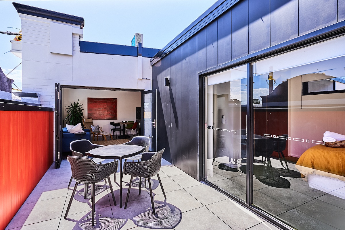 A spacious outdoor terrace features a round table surrounded by four chairs, with sliding glass doors leading to the interior. A vibrant yellow bed is visible through the glass, while the modern architecture is complemented by a clear blue sky.