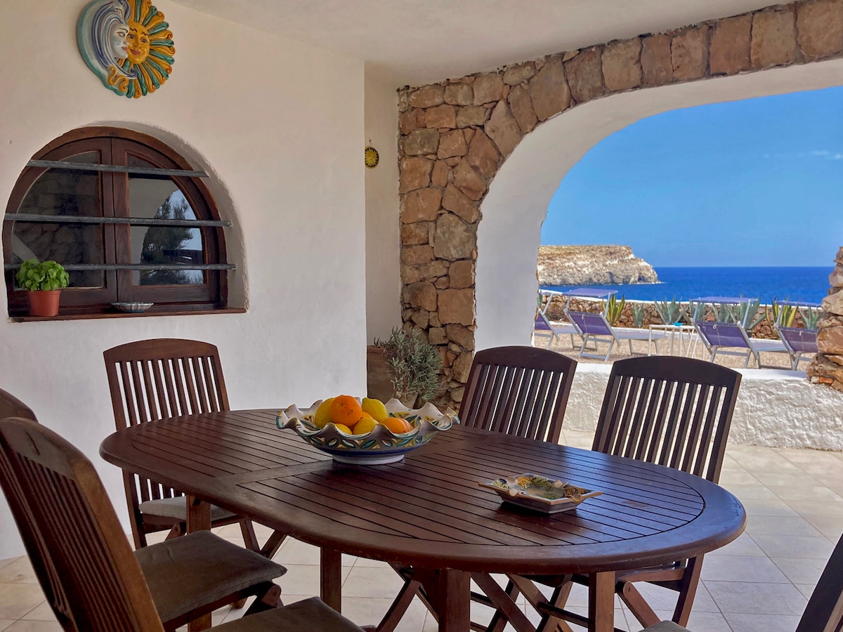 A wooden dining table with matching chairs is set under an arched stone entrance, offering a view of the sea. A bowl of fresh fruit sits at the center of the table. Sun loungers are visible in the background against a blue ocean backdrop.